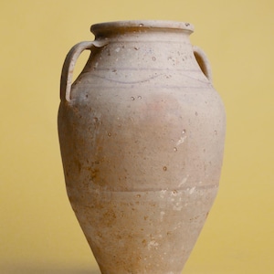 Puede incluir: Un jarrón grande de cerámica beige con dos asas. El jarrón tiene un cuerpo redondeado y un labio ligeramente acampanado.