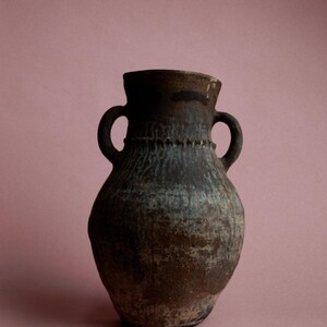 May include: A rustic, brown ceramic vase with two handles. The vase has a textured surface and a slightly flared rim. The color is a mix of brown and gray, with a hint of blue. The vase is set against a pink background.