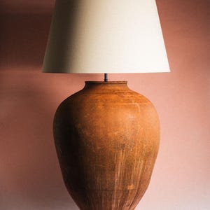 May include: A table lamp featuring a large, bulbous, terracotta-coloured clay vessel as its base. The base has a weathered, textured surface with vertical streaking. It is topped with a simple, off-white, conical fabric lampshade.