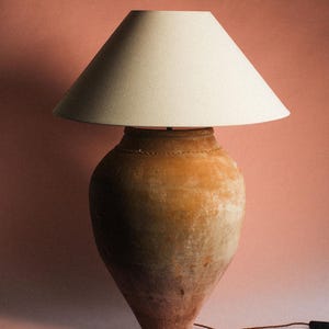Pode incluir: Um candeeiro de mesa com uma base de vaso de terracota desgastada, caracterizada por uma forma arredondada e cónica em tons de castanho terroso. O candeeiro é rematado com um abajur cónico largo de tecido em tom branco sujo e um fio fino.