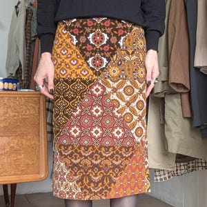 Può includere: Una gonna midi a fantasia patchwork nei toni del marrone, arancione e giallo. La gonna presenta vari motivi geometrici e floreali. La persona indossa una maglia nera a maniche lunghe e scarpe nere.