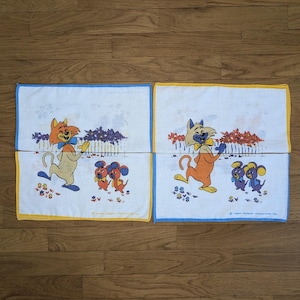 Peut inclure: Deux mouchoirs en tissu blanc avec une bordure jaune. Chaque mouchoir représente un chat de dessin animé et deux souris. Le chat est orange et blanc avec un nœud papillon bleu. Les souris sont violettes et blanches. Le texte "TWENTY BROTHERS INDUSTRIAL INC." est imprimé dans le coin inférieur droit d'un mouchoir.