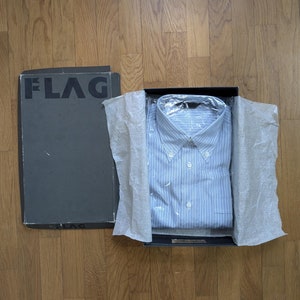 Può includere: Una camicia a righe blu e bianche con bottoni è piegata e posta all'interno di una scatola nera con la scritta "FLAG" stampata sul coperchio. La scatola è appoggiata su una superficie di legno.