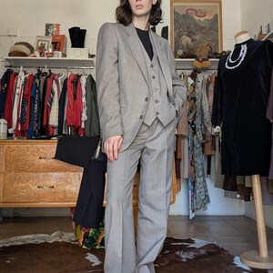 Puede incluir: Un traje de tres piezas gris claro con blazer, chaleco y pantalón de pernera ancha. El traje se lleva con un top negro y zapatos rojos. El fondo incluye percheros con ropa y una cómoda de madera.