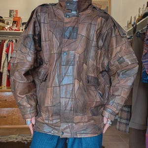 Può includere: Giacca in pelle marrone con design patchwork. La giacca ha un collo alto, chiusure con bottoni e più tasche. La giacca è di colore marrone medio e ha un'estetica vintage.