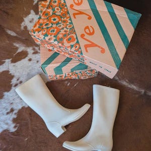 Könnte beinhalten: Ein Paar einfarbige, weiße wadenhohe Stiefel mit niedrigem Blockabsatz. Die Stiefel werden von einem Karton begleitet, der auf einer Seite ein sich wiederholendes orange-weißes Blumenmuster und auf der anderen türkise Diagonalstreifen zeigt.