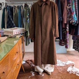 Può includere: Un lungo cappotto marrone con chiusura a bottoni e collo. Il cappotto è realizzato in tessuto strutturato e arriva al ginocchio. La persona indossa pantaloni color crema e scarpe fantasia. Appendiabiti sono visibili sullo sfondo.