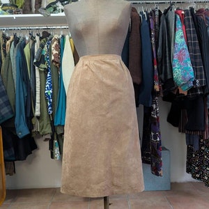 Può includere: Una gonna di camoscio marrone su un manichino in un negozio di abbigliamento. La gonna è lunga fino al ginocchio e ha una silhouette dritta. Il manichino è in piedi su un tappeto di pelle di mucca bianca e marrone.