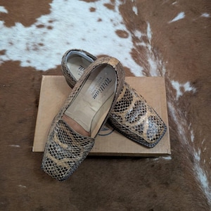 Peut inclure: Paire de mocassins à motif peau de serpent, à bout carré, dans des tons marron et beige, posés sur une boîte en carton. Les chaussures ont une étiquette visible à l'intérieur. Le fond est un tapis en peau de vache marron et blanc.
