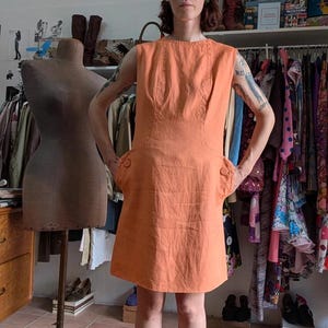 Può includere: Un abito arancione senza maniche con due tasche frontali e dettagli con bottoni. L'abito è lungo fino al ginocchio e sembra essere realizzato in un tessuto strutturato. Un manichino vintage è a sinistra e un attaccapanni sullo sfondo.