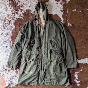 Op de afbeelding: Een groene capuchonjas met een bruine imitatiebontvoering. De jas heeft meerdere zakken en een riem.