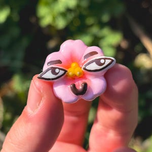 Puede incluir: Un objeto rosa con forma de flor con ojos de dibujos animados y una boca sonriente. La flor tiene un centro amarillo y se sostiene en una mano sobre un fondo verde borroso.