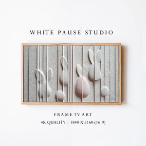 May include: A framed wall art piece featuring a textured gray background with vertical lines and raised bunny and egg shapes in white and pink. The frame is made of light-colored wood. The text "WHITE PAUSE STUDIO" and "FRAME TV ART" are visible.