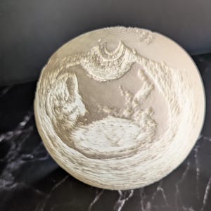 Könnte beinhalten: Ein kreisförmiges, cremefarbenes Objekt mit strukturierter Oberfläche. Das Objekt weist ein erhabenes, detailliertes Design auf, möglicherweise eine Landschaft oder topografische Darstellung. Die Gesamtästhetik ist monochrom, mit subtilen Farbtonvariationen.