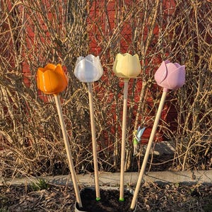 Può includere: Quattro picchetti da giardino a forma di tulipano colorati in una fioriera grigia. Le teste dei tulipani sono arancioni, bianche, gialle e rosa, ciascuna attaccata a un tassello di legno. I picchetti sono piantati in terra scura, con una statuina di fata visibile.