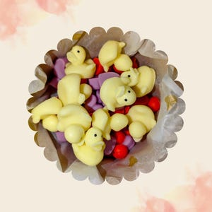 Könnte beinhalten: Nahaufnahme eines dekorativen Papierbechers gefüllt mit gelben Enten- und Schweinchen-förmigen Süßigkeiten, lila herzförmigen Süßigkeiten und roten runden Süßigkeiten. Der Becher hat einen gewellten Rand und ist mit Pergamentpapier ausgekleidet.