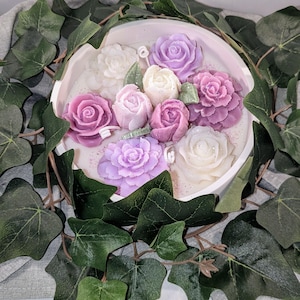 May include: A white circular candle with a floral design. The candle features multiple pink and purple roses with green leaves. The candle is surrounded by a garland of green ivy.