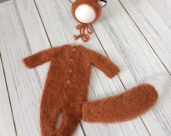 May include: A brown knitted fox costume for a newborn baby. The costume includes a hat with ears, a jumpsuit, and a tail.