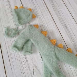 May include: A green knitted baby dinosaur costume with a matching hat. The dinosaur has yellow spikes on its back and tail. The hat has yellow spikes on top.