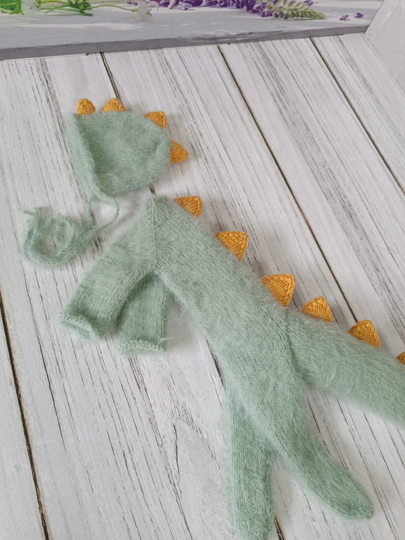 May include: A green knitted baby dinosaur costume with a matching hat. The dinosaur has yellow spikes on its back and tail. The hat has yellow spikes on top.