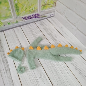 May include: A green knitted baby dinosaur costume with a matching hat. The costume has yellow spikes and a fuzzy texture.