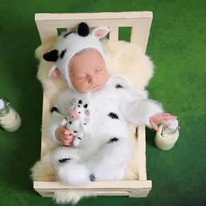 May include: A newborn baby wearing a white and black cow costume sleeps in a white wooden crib with a white furry blanket. The baby is holding a small stuffed cow. Two glass bottles of milk are on either side of the crib.