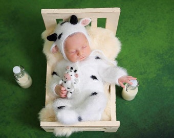 May include: A newborn baby wearing a white and black cow costume sleeps in a white wooden crib with a white furry blanket. The baby is holding a small stuffed cow. Two glass bottles of milk are on either side of the crib.