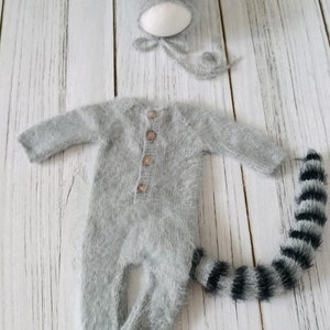 May include: A gray knitted baby outfit with a raccoon tail and a matching raccoon hat. The outfit has a button closure and the hat has black and white stripes.