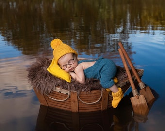 Puede incluir: Un bebé con un gorro de punto amarillo y gafas duerme en un bote de madera con remos. El bote flota en un cuerpo de agua tranquilo.