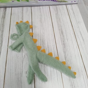 May include: A green knitted dinosaur costume with yellow accents. The costume has a hood and a long tail.