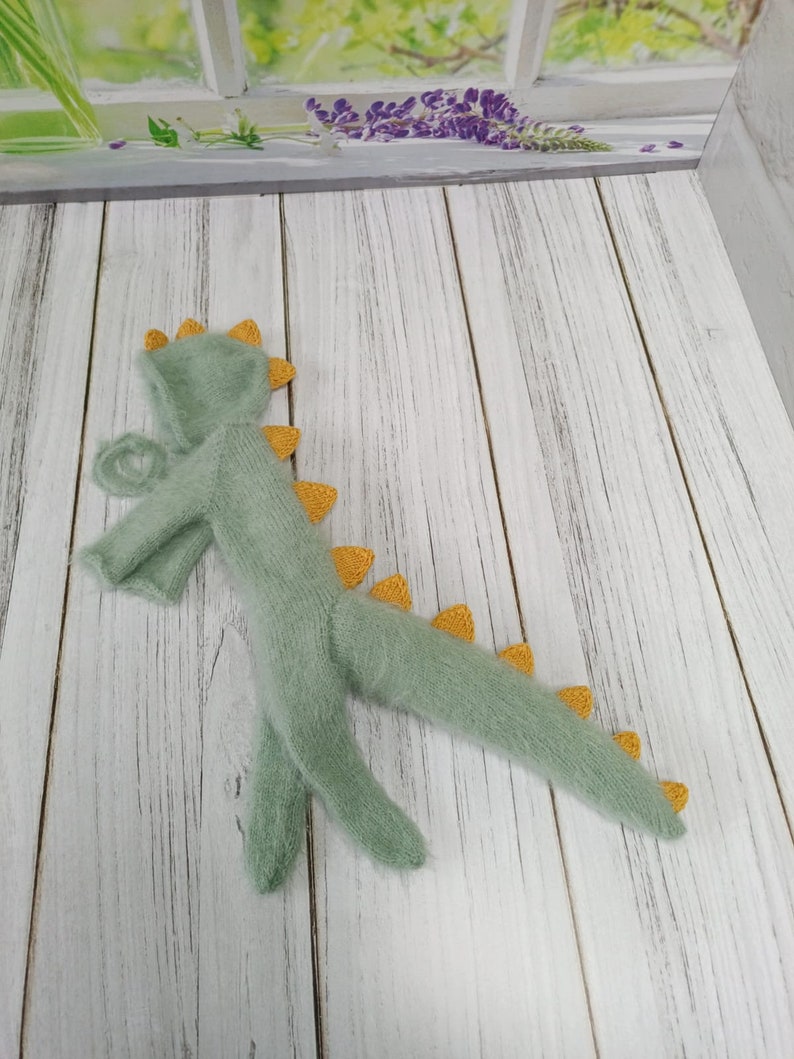 May include: A green knitted dinosaur costume with yellow accents. The costume has a hood and a long tail.
