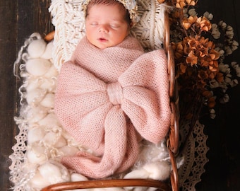 May include: A newborn baby girl is sleeping in a small wooden crib, wrapped in a pink knitted blanket with a large bow. The crib is decorated with white fluffy yarn and a crocheted doily. The baby is wearing a white flower crown.