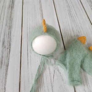 May include: A green knitted baby hat and matching sweater with yellow dinosaur horns. The hat is on a white styrofoam head and the sweater is lying on a white wooden surface.