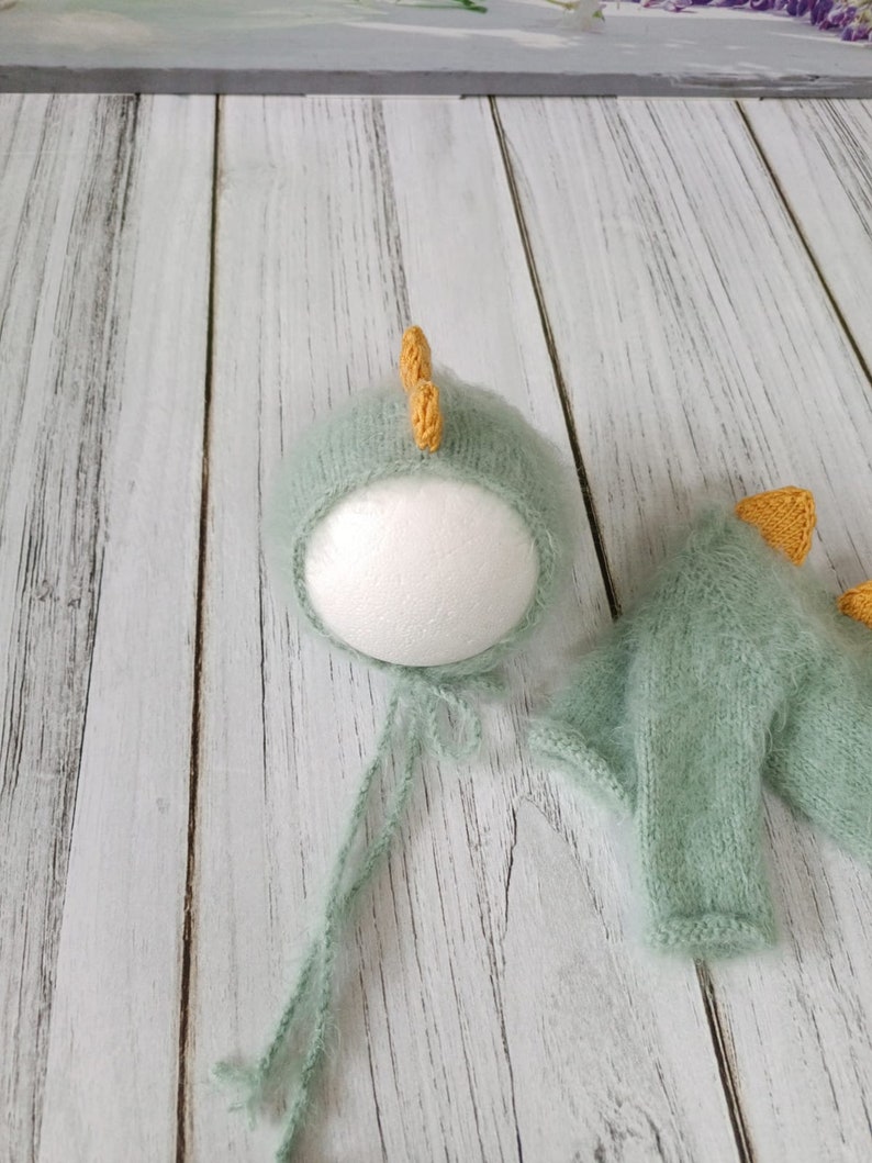 May include: A green knitted baby hat and matching sweater with yellow dinosaur horns. The hat is on a white styrofoam head and the sweater is lying on a white wooden surface.