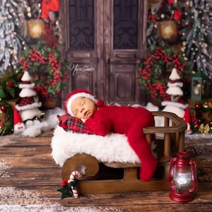 May include: A newborn baby wearing a red Santa Claus outfit and hat, sleeping on a sled with a white fur blanket and a plaid pillow. The sled is on a wooden surface, with Christmas decorations in the background.
