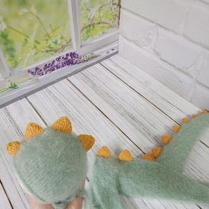 May include: A green and yellow knitted dinosaur costume for a newborn baby. The costume includes a hat and a onesie with a tail.