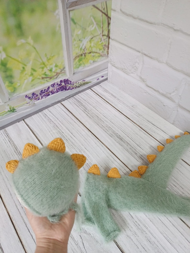 May include: A green and yellow knitted dinosaur costume for a newborn baby. The costume includes a hat and a onesie with a tail.