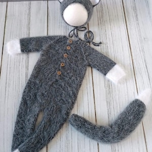 May include: Gray knitted baby outfit with a cat theme. The outfit includes a hooded hat with cat ears, a long-sleeved jumpsuit, and a tail.