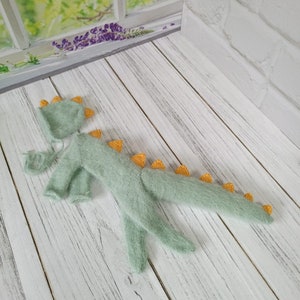 May include: A light green knitted baby dinosaur costume with yellow spikes. The costume includes a hat and a onesie.