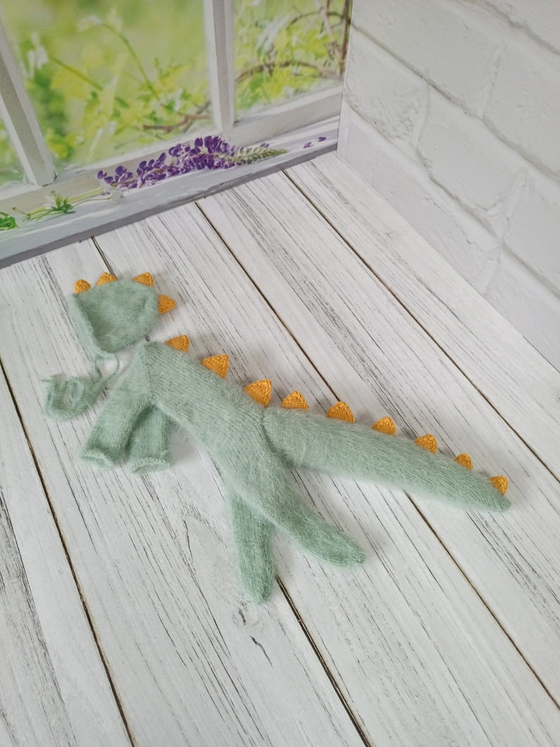 May include: A light green knitted baby dinosaur costume with yellow spikes. The costume includes a hat and a onesie.