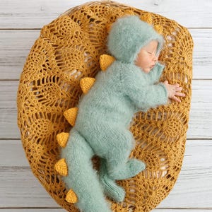 May include: A baby wearing a light blue knitted dinosaur costume with yellow accents is sleeping on a yellow crocheted blanket.