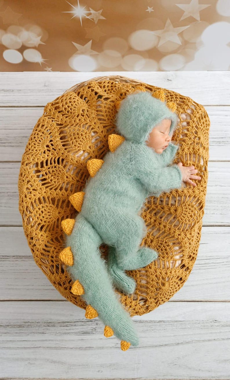 May include: A baby wearing a light blue knitted dinosaur costume with yellow accents is sleeping on a yellow crocheted blanket.
