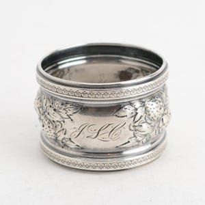 May include: A cylindrical silver napkin ring featuring an embossed strawberry and leaf motif around the exterior. The center of the band displays an engraved monogram with the letters JLC in a cursive script, framed by decorative borders.