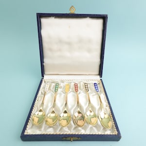 May include: A set of six gold-toned metal spoons with colorful enamel accents on the handles, presented in a dark blue rectangular box with a white satin-lined interior. Each spoon features a unique colored enamel inlay at the top.