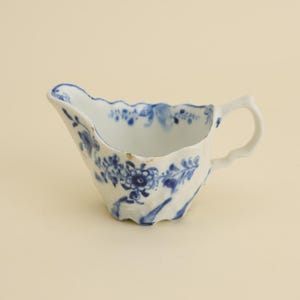 May include: A white porcelain gravy boat with a blue floral pattern. The boat has a scalloped rim and a curved handle. The blue floral design is hand-painted and covers the exterior of the boat.
