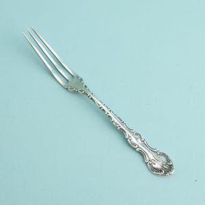 May include: A silver-toned metal fork featuring three slender tines and an ornate, embossed handle with intricate scrollwork and curved detailing. The metal surface has a polished finish, reflecting light along the decorative stem.