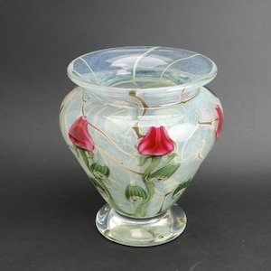 Può includere: Un vaso decorativo in vetro con sfondo azzurro chiaro e accenti floreali rossi. Il vaso ha una forma bulbosa con un bordo svasato e una base trasparente e arrotondata. I fiori sono raffigurati in modo stilizzato, aggiungendo un tocco di eleganza.