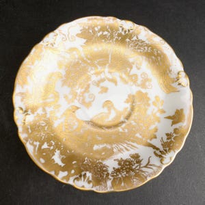 Pode incluir: Um pires redondo de porcelana branca com um padrão metálico dourado intrincado de pássaros, folhagem e motivos florais. O pires tem uma borda recortada e uma reentrância circular central, com o design dourado cobrindo a superfície.