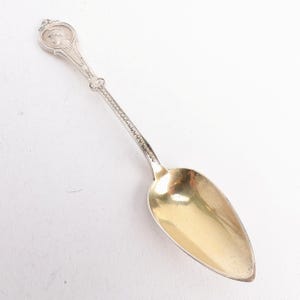 May include: A silver spoon with a gold-colored bowl. The handle features a detailed design with a circular emblem at the top. The spoon's handle is adorned with intricate patterns and a decorative ring.