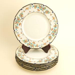 May include: A set of white ceramic plates with a floral design. The plates have a scalloped edge with a blue and gold trim. The floral pattern includes orange, blue, and yellow flowers. One plate is displayed on a wooden stand, with several stacked below.